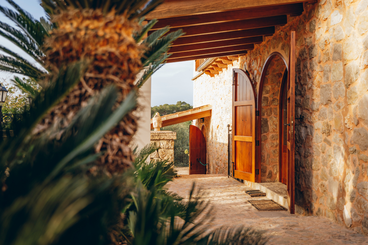 Blick auf eine Steinmauer mit Holzladen und offener Tür, umgeben von Palmen im Sonnenlicht.