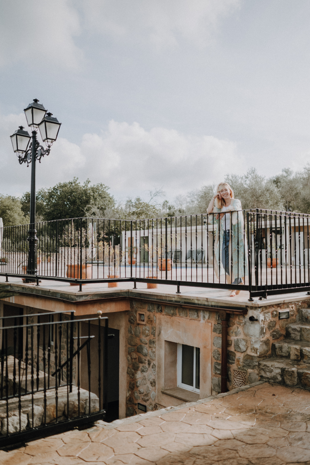 Eine lachende Frau steht auf einer Terrasse mit Steinboden und Metallgeländern, im Hintergrund sind Bäume und ein Gebäude sichtbar.