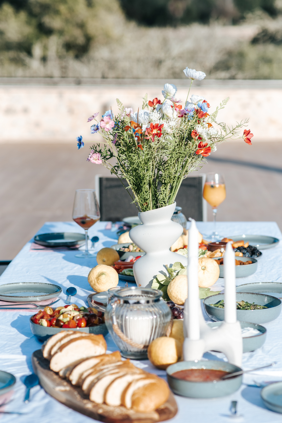 Ein festlich gedeckter Tisch mit Blumenstrauß in einer weißen Vase, Kerzen, verschiedenen Speisen und Getränken, im Freien bei Sonnenlicht.