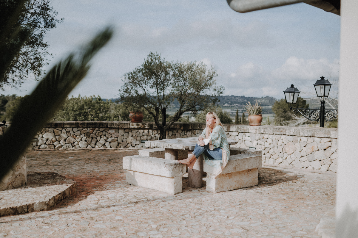 Frau sitzt auf einer Steinbank im Freien, umgeben von einer Steinmauer und Pflanzen, mit Blick auf eine Landschaft und bewölkten Himmel.