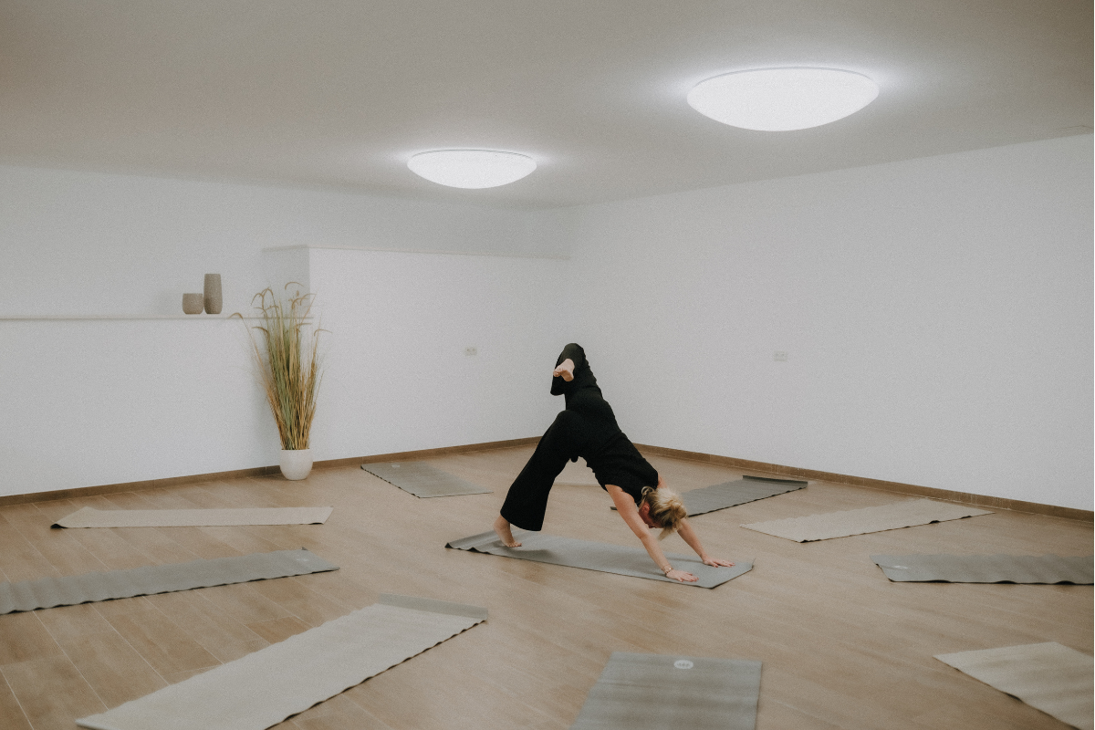 Eine Frau macht Yoga in einem hellen, leeren Raum mit Holzfußboden. Sie befindet sich in einer vorwärts gerichteten Hund-Position auf einer Yogamatte, umgeben von mehreren liegen gelassenen Matten. Es gibt eine Vase mit langen Gräsern und zwei dekora