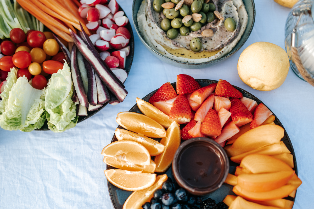Bunte Frucht- und Gemüseschale mit Erdbeeren, Orangen, Honigmelone, Heidelbeeren, Radieschen, Cherry-Tomaten, Salatblättern, Karotten, Gurken, Zitronen, sowie Dips und eine Wasserflasche auf einem blauen Tischtuch.