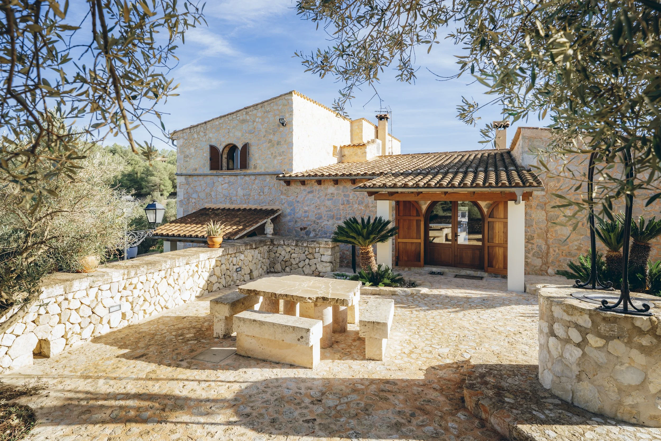 Ein mediterranes Haus mit Steinmauern, Holztüren und Terrassenmöbeln, umgeben von Olivenbäumen und Palmen, bei sonnigem Himmel.