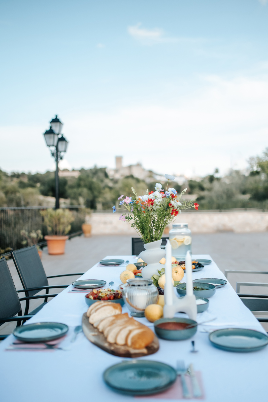 Ein gedeckter Esstisch im Freien mit einer weißen Tischdecke, einer Vase mit bunten Blumen, Kerzen, Brot, Obst und Geschirr, auf einem Balkon mit Blick auf eine Landschaft mit Bäumen und Häusern.