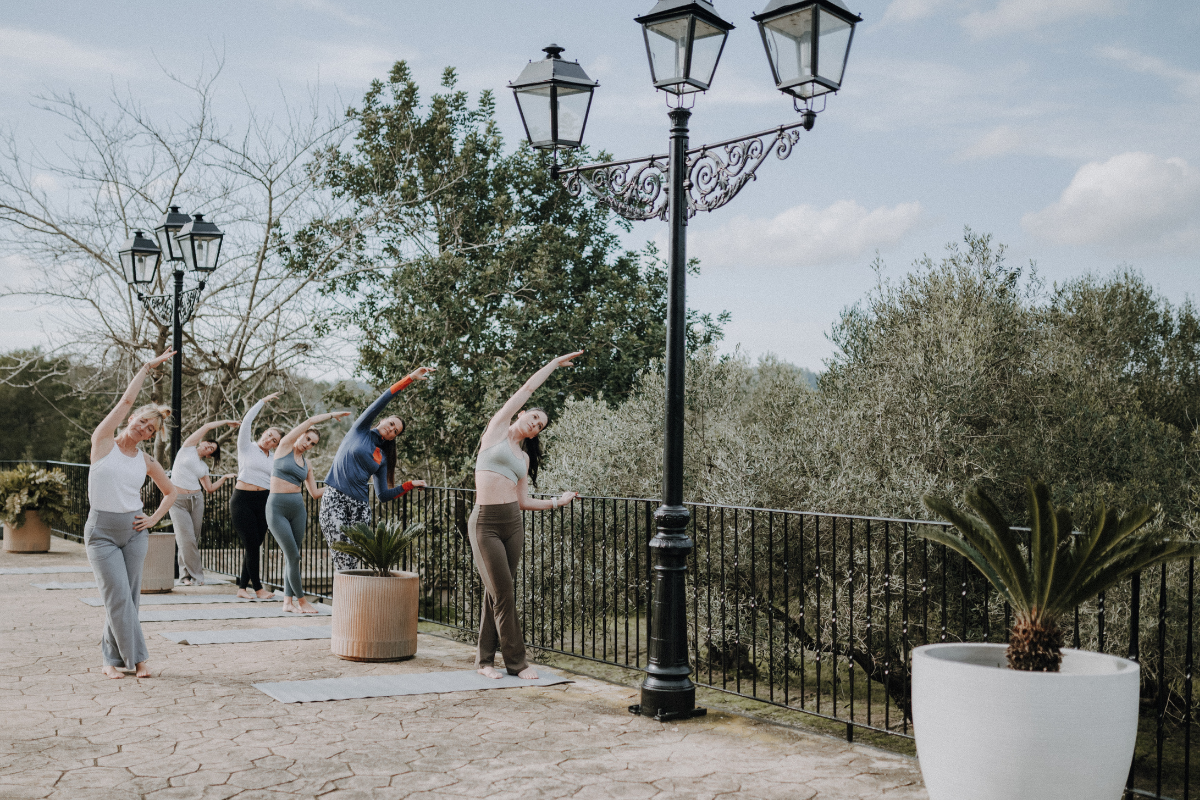 Frauen, die Yoga auf einer Terrasse im Freien machen, während sie Dehnübungen im Stehen ausführen, bei Sonnenlicht, neben einer Straßenlaterne und großen Pflanzkübeln mit Pflanzen.