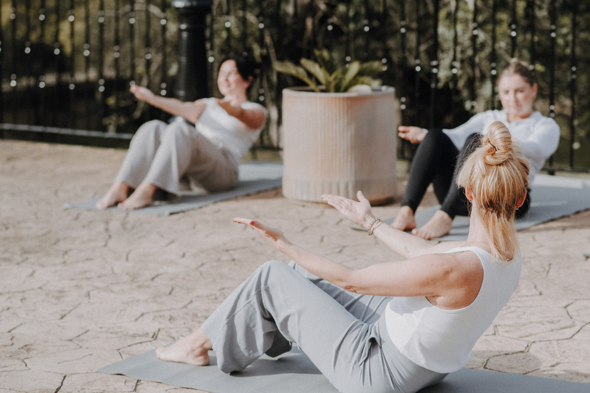 Drei Frauen machen Yoga im Freien auf einer Terrasse, die Frauen im Vordergrund hat blonde Haare im Dutt und trägt ein weißes Top, sie führen eine Sit-up-Übung aus, im Hintergrund sind weitere Frauen, die ebenfalls Yoga auf Decken praktizieren.