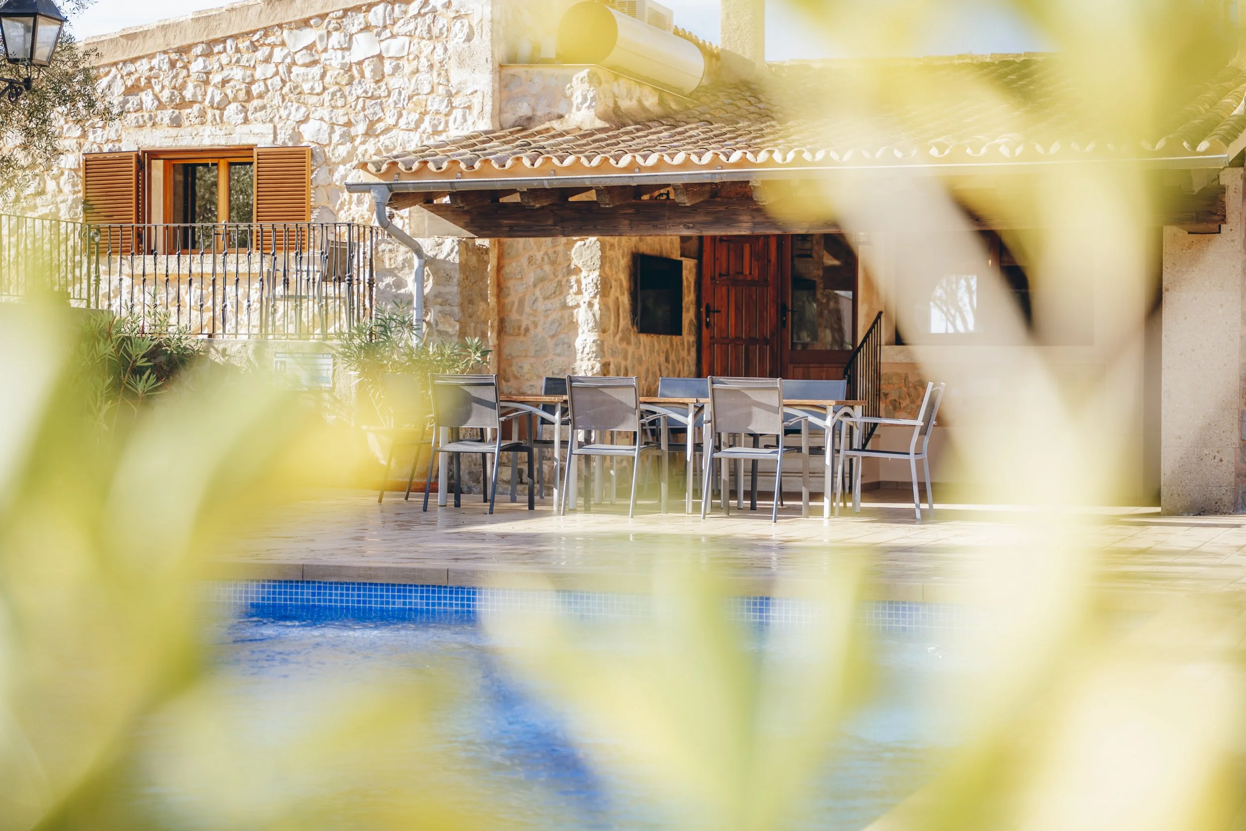 Urige Steinhütte mit Terrassenmöbeln, Blick durch Blätter auf einen Pool im Vordergrund, sonniges Wetter, ländliche Umgebung.
