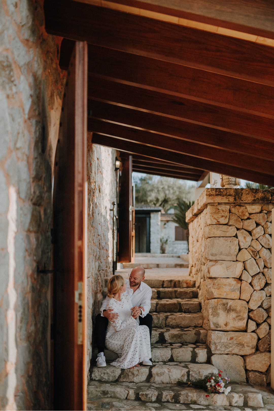Ein Mann und eine Frau sitzen auf Steinstufen vor einem Haus aus Naturstein, umgeben von Steinstufen, während Blumen vor ihnen liegen. Sie lachen und schauen sich liebevoll an.