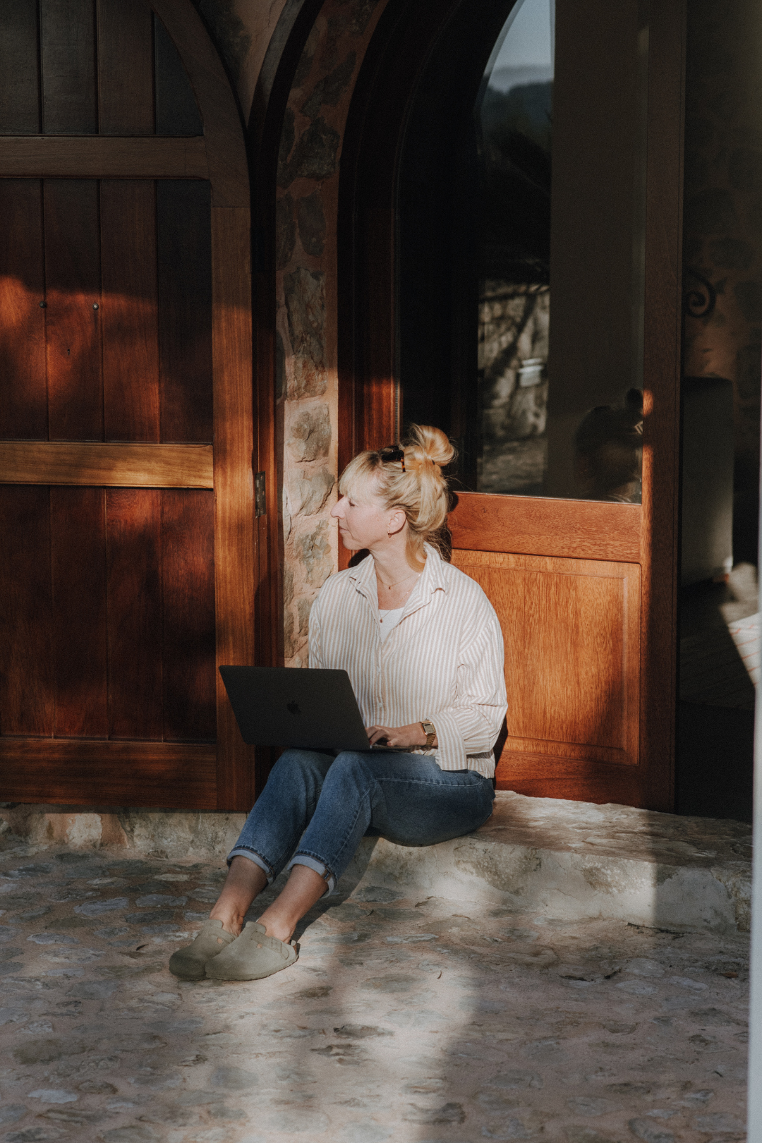 Frau sitzt mit einem Laptop vor einer holzverzierten Tür im Sonnenlicht, an einer Steinmauer.