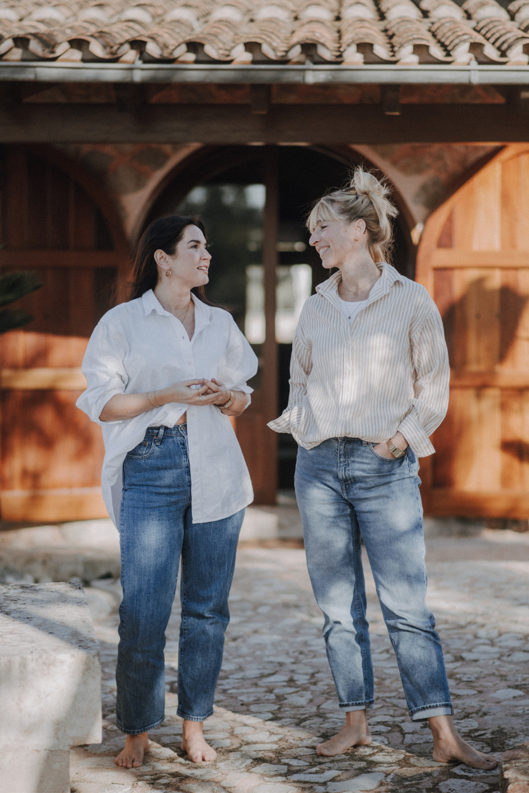 Zwei Frauen stehen barfuß auf einem gepflasterten Weg, sprechen und lachen miteinander vor einem Holzgebäude mit einem Schindeldach.