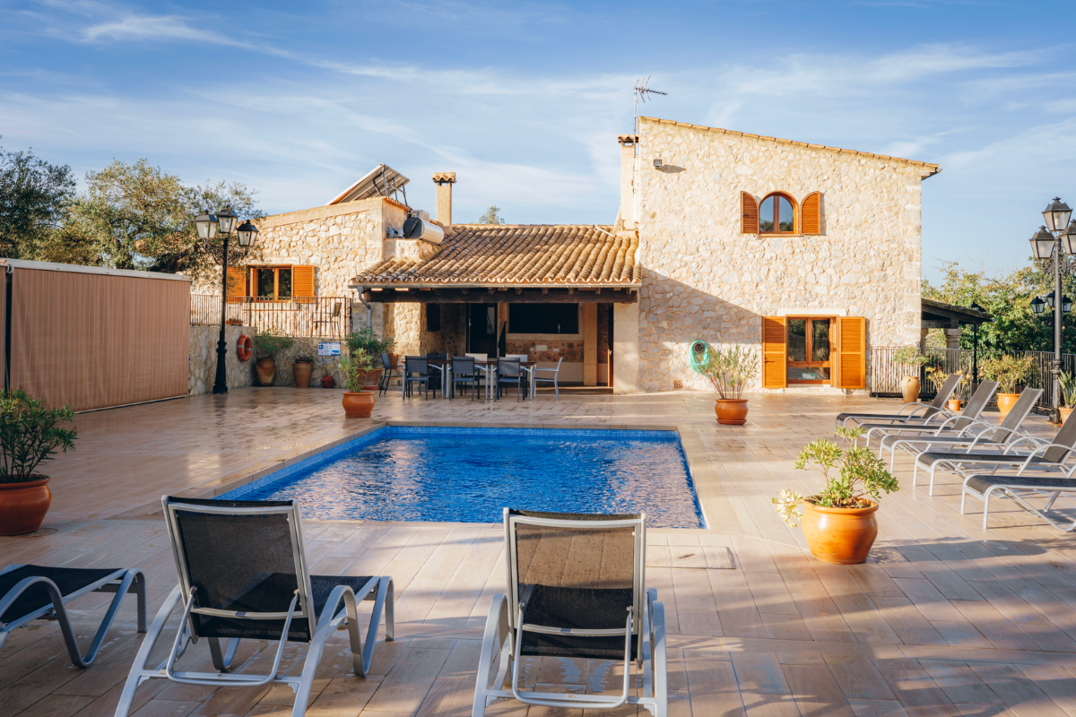Ein Außenpool im Innenhof eines Hauses mit Sonnenschirmen und Liegestühlen, umgeben von Terrakottatöpfen mit Pflanzen, vor einem Steinhaus mit Fensterläden. Es ist ein sonniger Tag mit blauem Himmel.