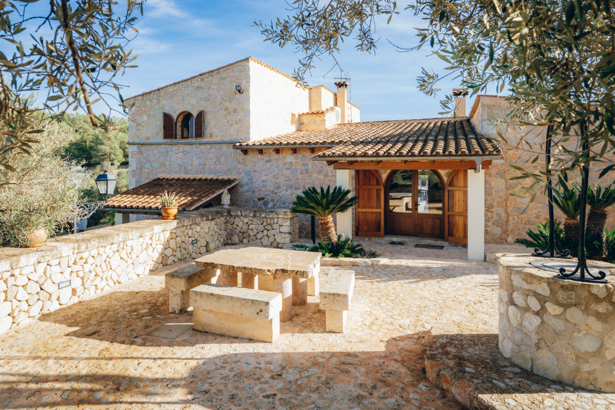 Ein traditionelles Haus mit Steinfassade, Terrakotta-Dachziegeln und Holzfenstern, umgeben von mediterranen Pflanzen, befindet sich auf einem steinigen Hof mit einem Steintisch und Bänken in der Sonne.