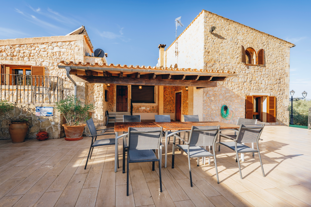 Ein Außenbereich eines Hauses mit Steinfassade, Tischen und Stühlen, Pflanzen in Terrakottatöpfen und Sonnenschirmen unter blauem Himmel.