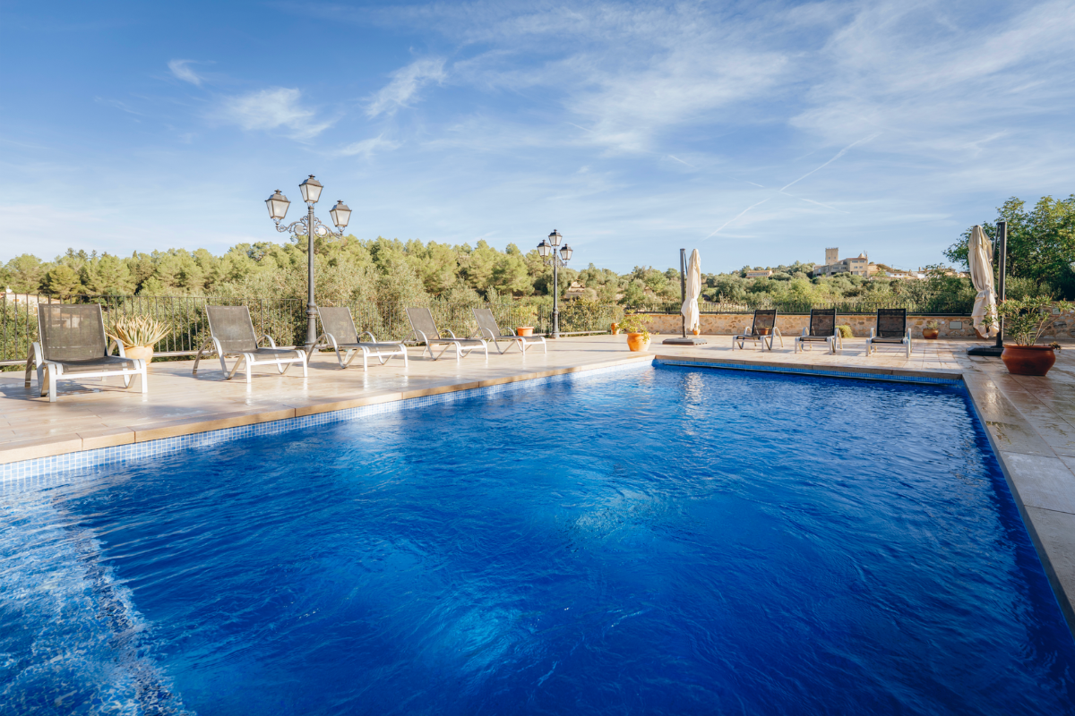 Ein Swimmingpool im Freien mit Sonnenliegen, Sonnenschirmen, Straßenlaternen und grünen Büschen im Hintergrund, unter einem blauen Himmel mit wenigen Wolken.