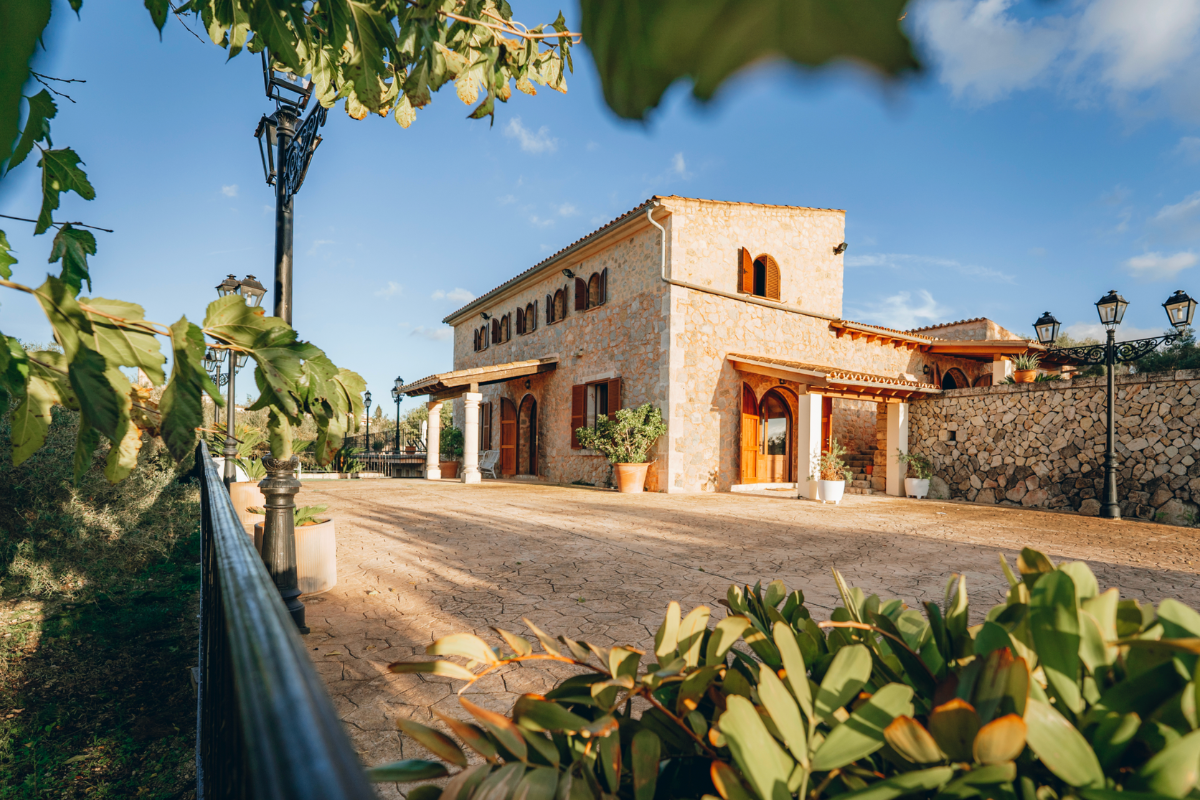 Ein Haus in einem mediterranen Stil mit Steinmauern und roten Türen, umgeben von einem Garten mit Pflanzen, unter einem blauen Himmel