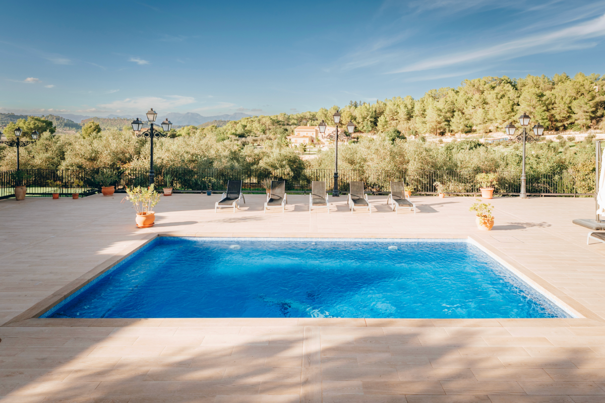 Ein Pool auf einer Terrasse mit Sonnenliegen und Lampen, umgeben von Bäumen und Bergen im Hintergrund bei sonnigem Himmel.