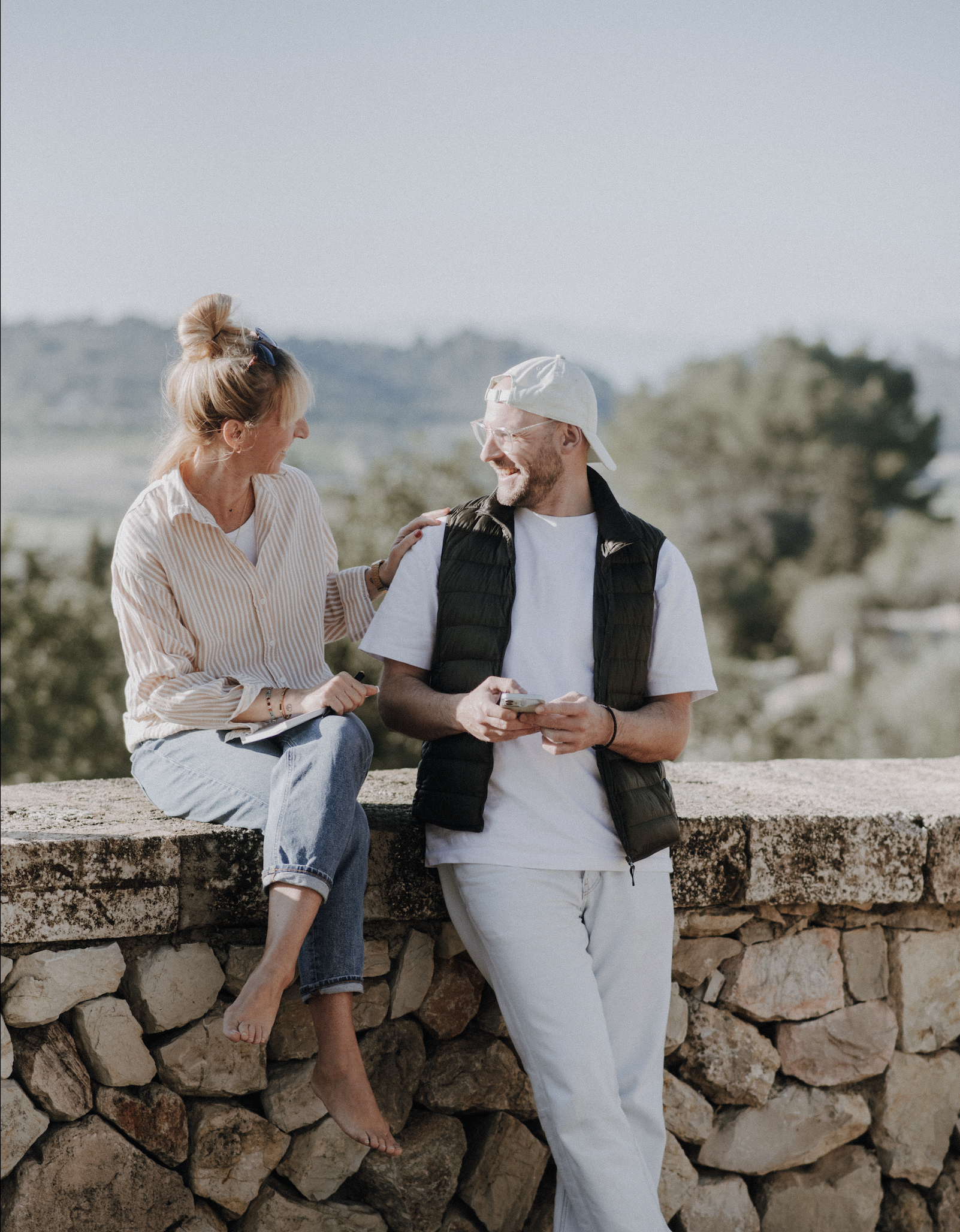Ein Mann und eine Frau sitzen und stehen auf einer Steinmauer im Freien, lachen und unterhalten sich. Der Mann trägt eine weiße Kappe und ein schwarzes gepolstertes Weste, die Frau trägt ein gestreiftes Hemd und Jeans. Im Hintergrund sind Bäume und eine hügelige Landschaft sichtbar.