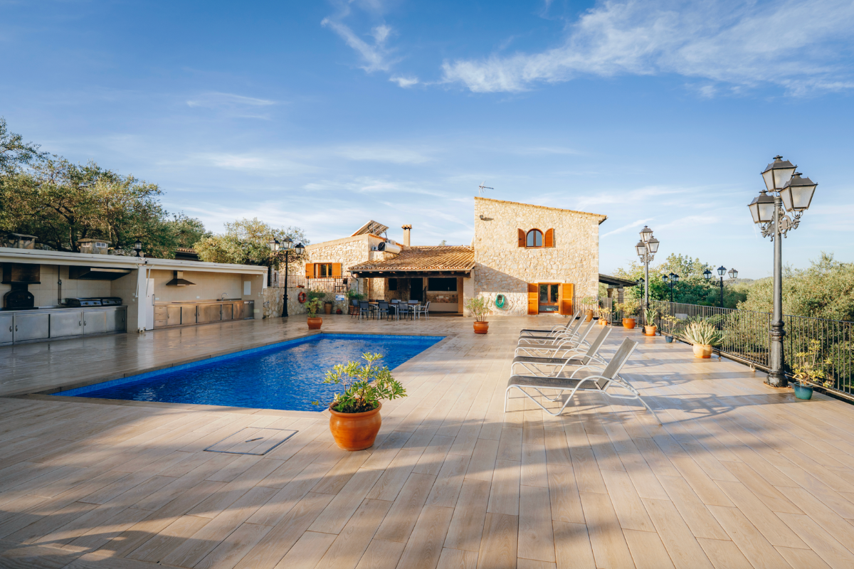 Ein Poolbereich im Freien mit Liegestühlen, Pflanzen in Töpfen, einem Gebäude im Hintergrund und Gartenlampen bei sonnigem Himmel.