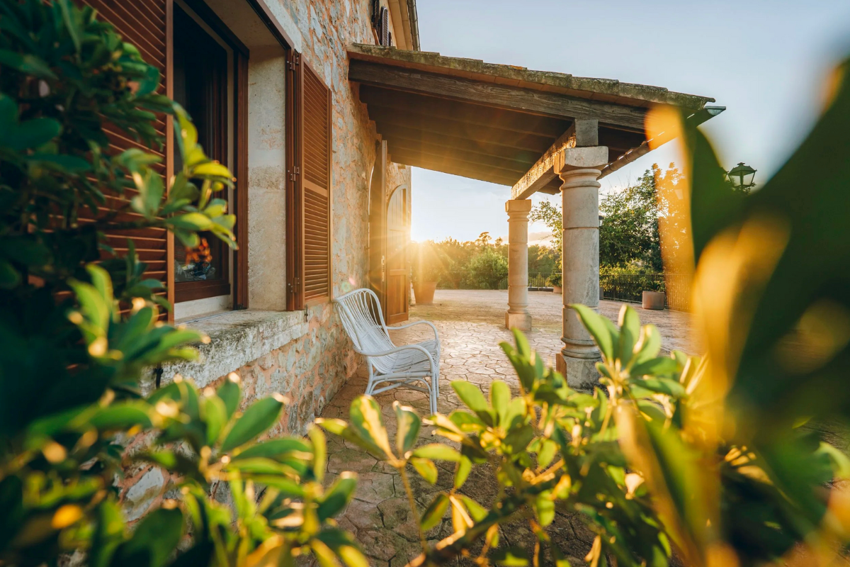 Sonnenuntergang hinter einem Haus mit einer Terrasse, weißen Stühlen, Holztüren, Steinmauern und alten Säulen, umgeben von grünen Pflanzen.