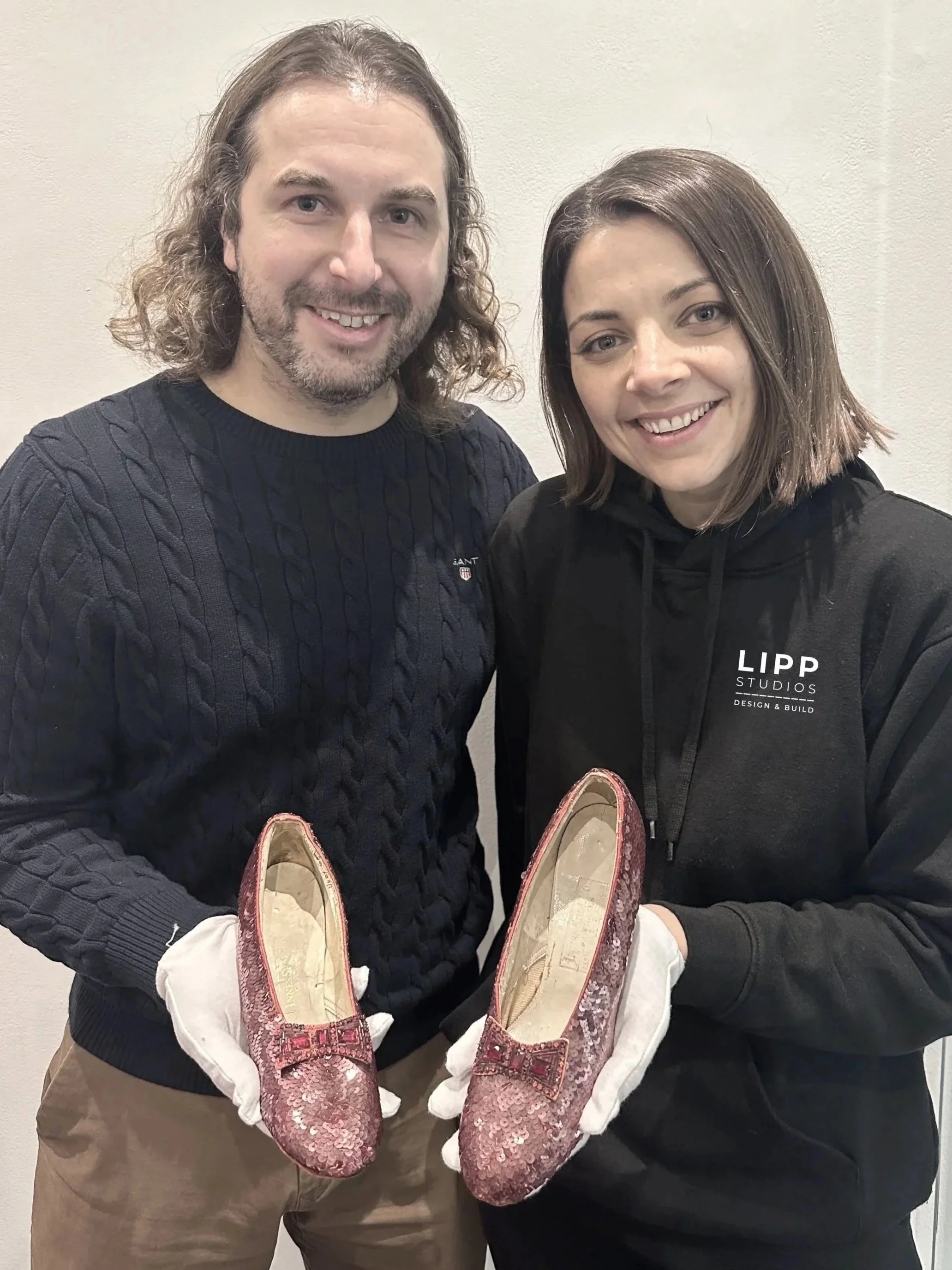 A man and woman smiling, holding pink glittery shoes with bows, standing against a gray wall.