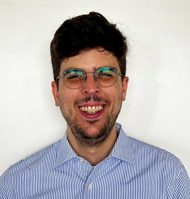 A man with glasses and a striped shirt smiling in front of a plain white background.
