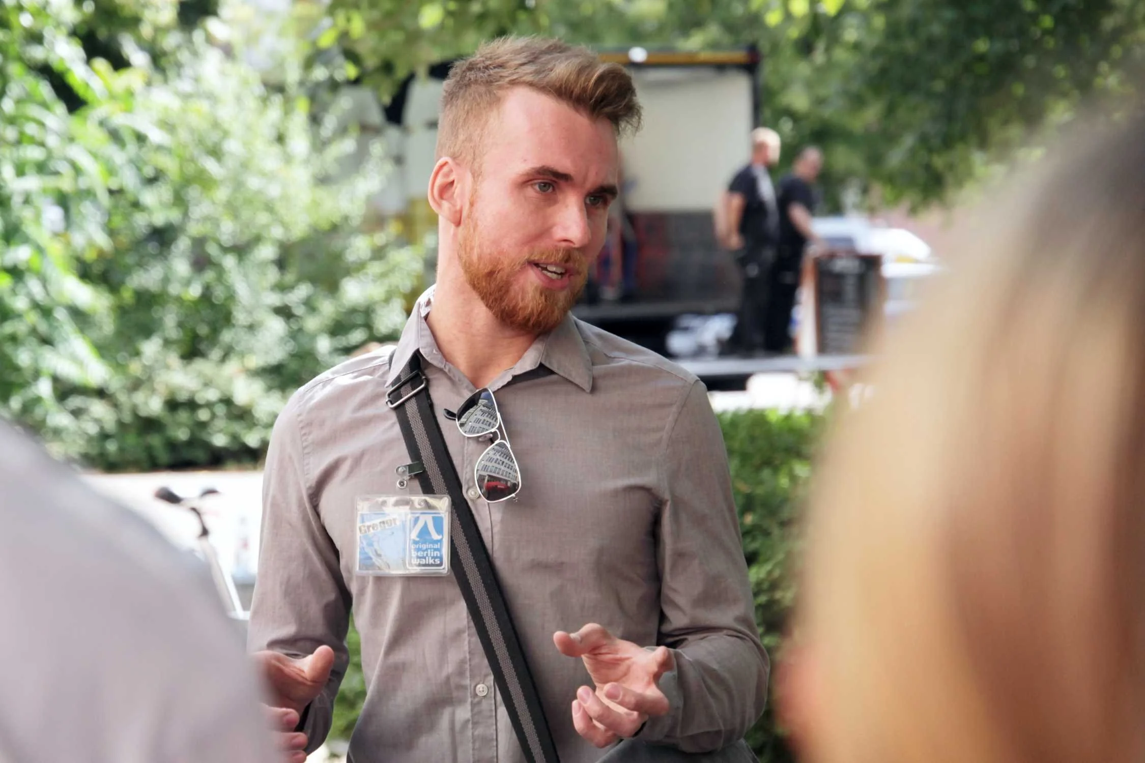 An Original Munich Walks Guide with light brown hair, a beard, wearing a gray shirt, sunglasses hanging from his collar, and a blue lanyard with a name badge, is talking to a group of people outdoors with green trees in the background.