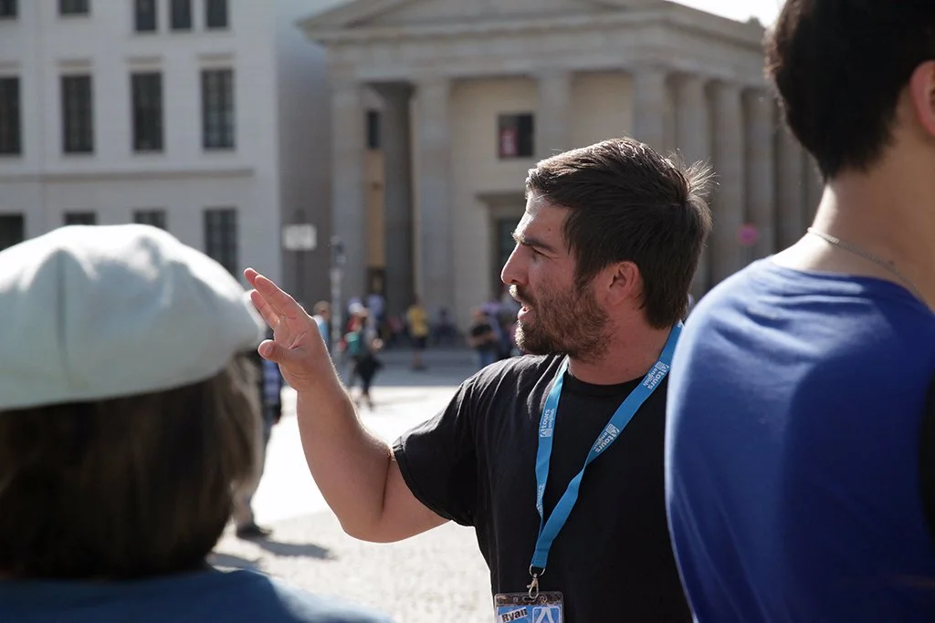 A man with short dark hair and a beard, wearing a black shirt and a blue lanyard, is gesturing with his right hand while talking to people on the street in front of a large building with columns.