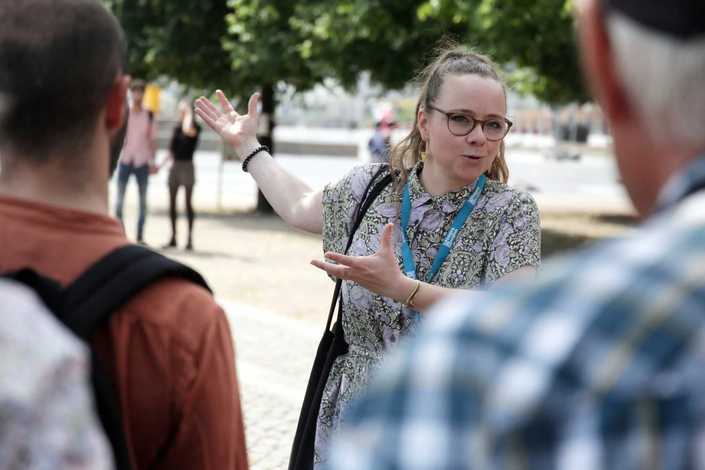 A woman with glasses and a patterned dress explaining something to a group of people outdoors. She is gesturing with her arms as she talks, and there are trees and blurred people in the background.