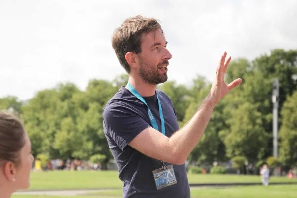 A man speaking outdoors, gesturing with his right hand, with a woman partially visible in the foreground, in a park with green trees in the background.