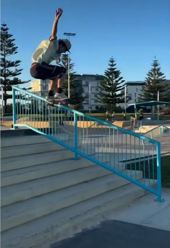 Skateboarder in mid-air executing a trick over a metal railing at a skate park, with stairs below, trees, and buildings in the background on a clear day.