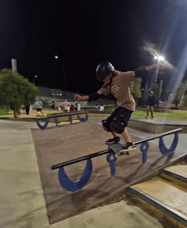 A young skateboarder wearing a helmet practicing a trick on a rail at a skatepark during the night. There are other people and large trees in the background, along with bright overhead lights illuminating the area.