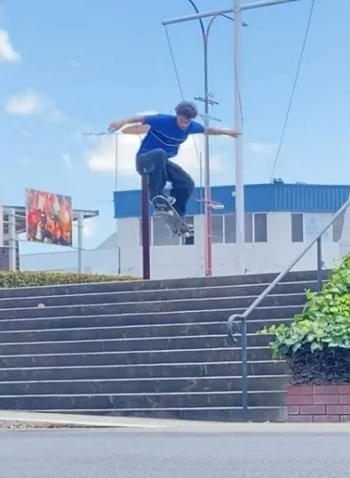A young man is skateboarding off a ledge on a city street, mid-air with arms outstretched, blue sky in the background.