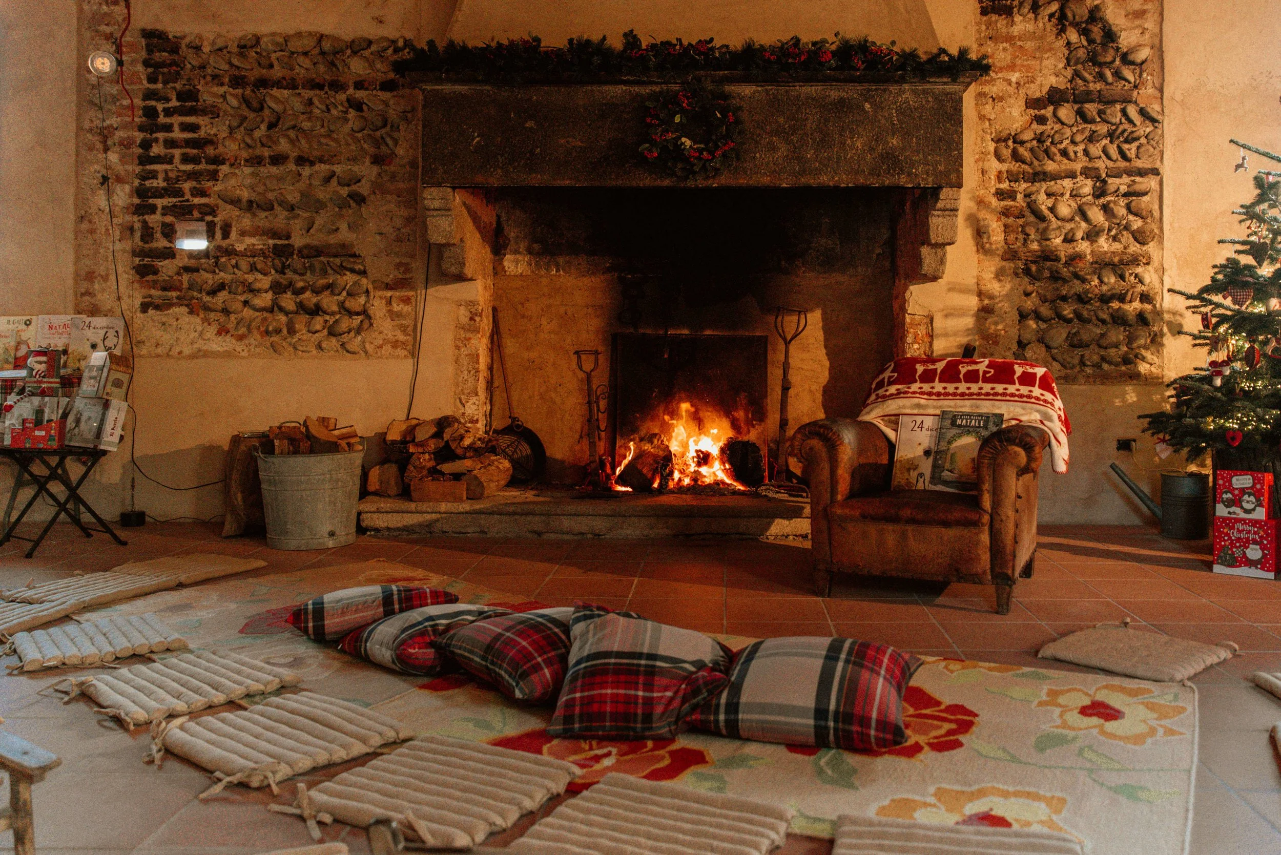Camino acceso con fuoco acceso, divano imbottito coperto con coperta natalizia, poltrona, albero di Natale decorato con luci e ornamenti, sedie con regalo di Natale, zona calda e accogliente. Piemonte – Castello di Cavagliano, Novara