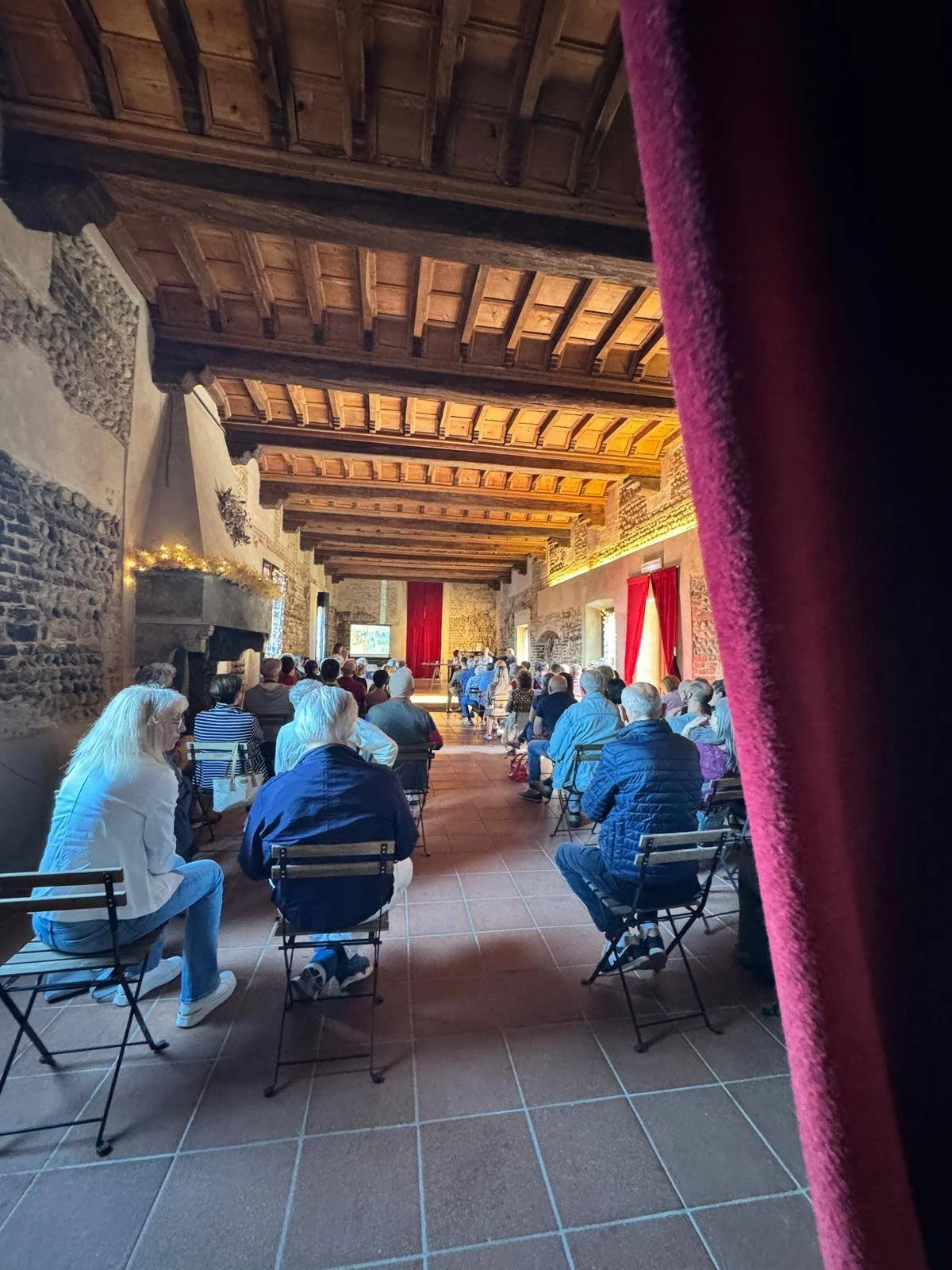 Persone sedute in un'aula con pareti di pietra e soffitto in legno, ascoltando una presentazione proiettata sullo schermo davanti a loro. Piemonte – Castello di Cavagliano, Novara