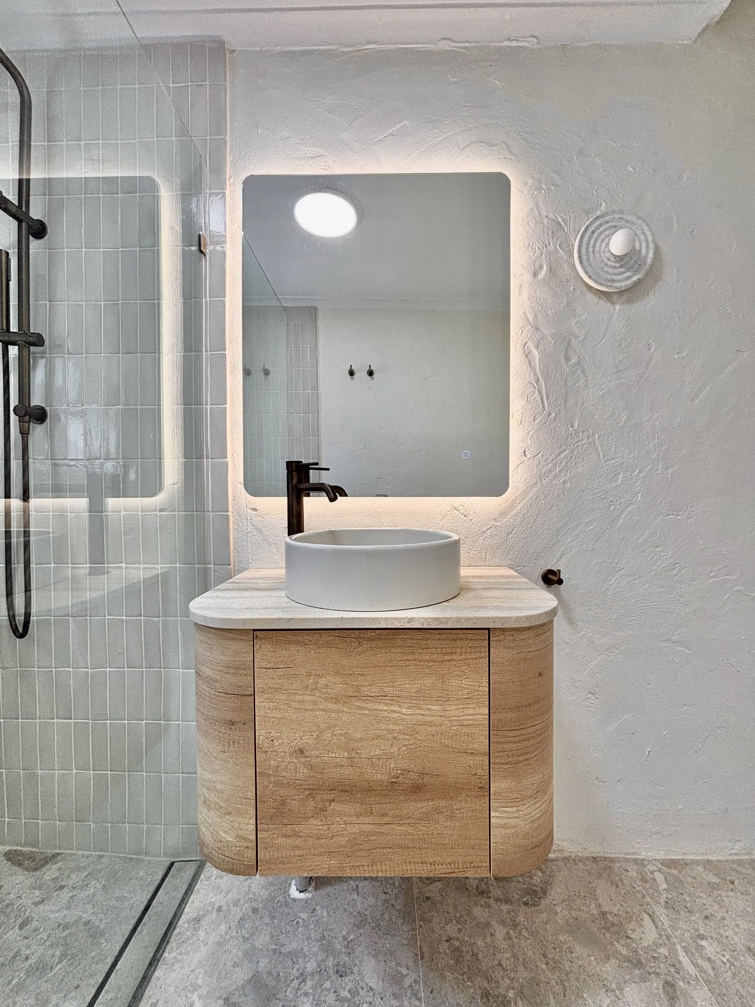 Modern bathroom with a round white vessel sink, black faucet, wooden vanity, mirror with backlit lighting, white textured wall, and shower area with glass door and beige tiles.