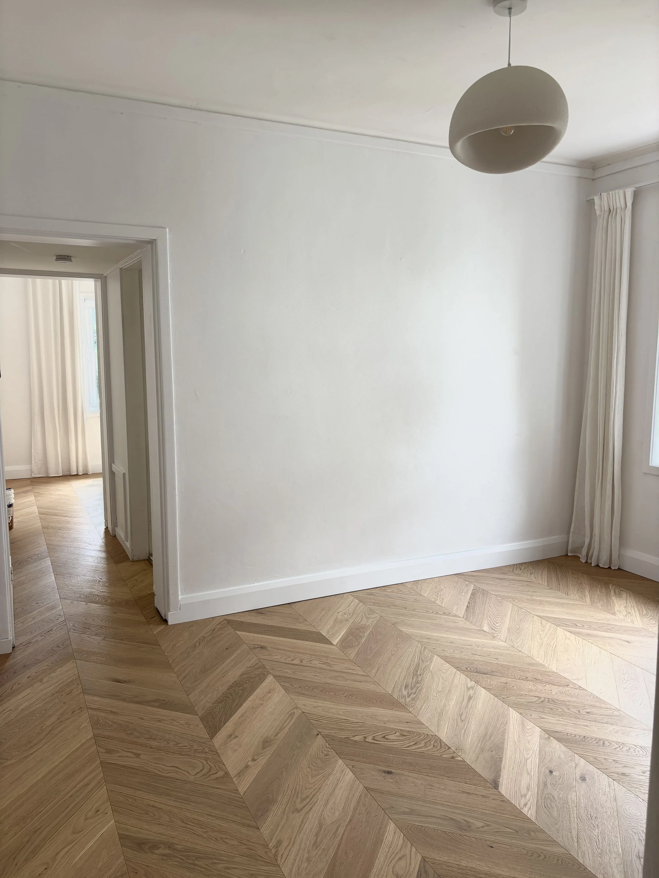 Empty room with white walls, wooden herringbone floor, and a ceiling light fixture.