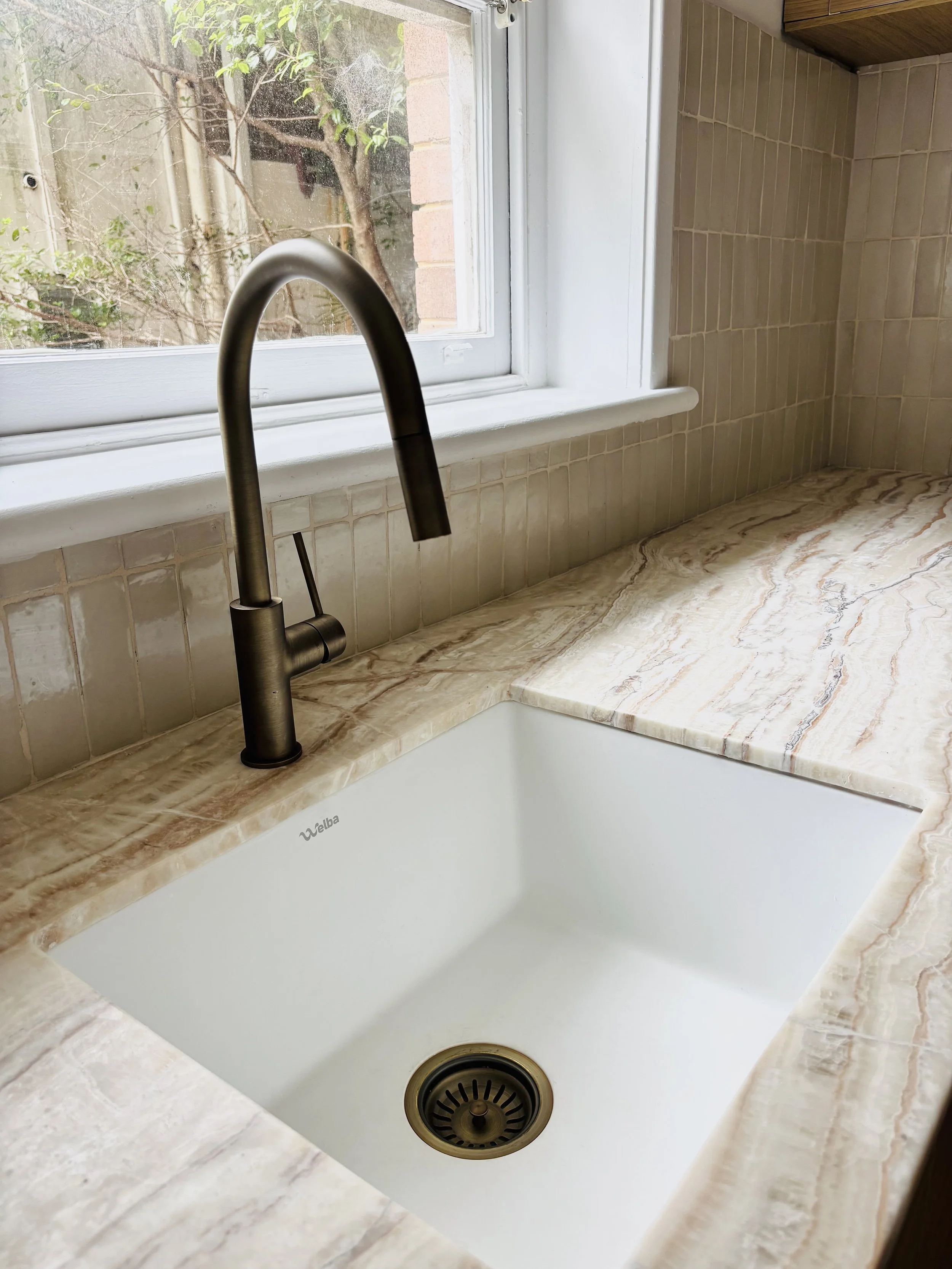 Kitchen sink below a window, with a black faucet and beige marble countertop, and a view of green trees outside.