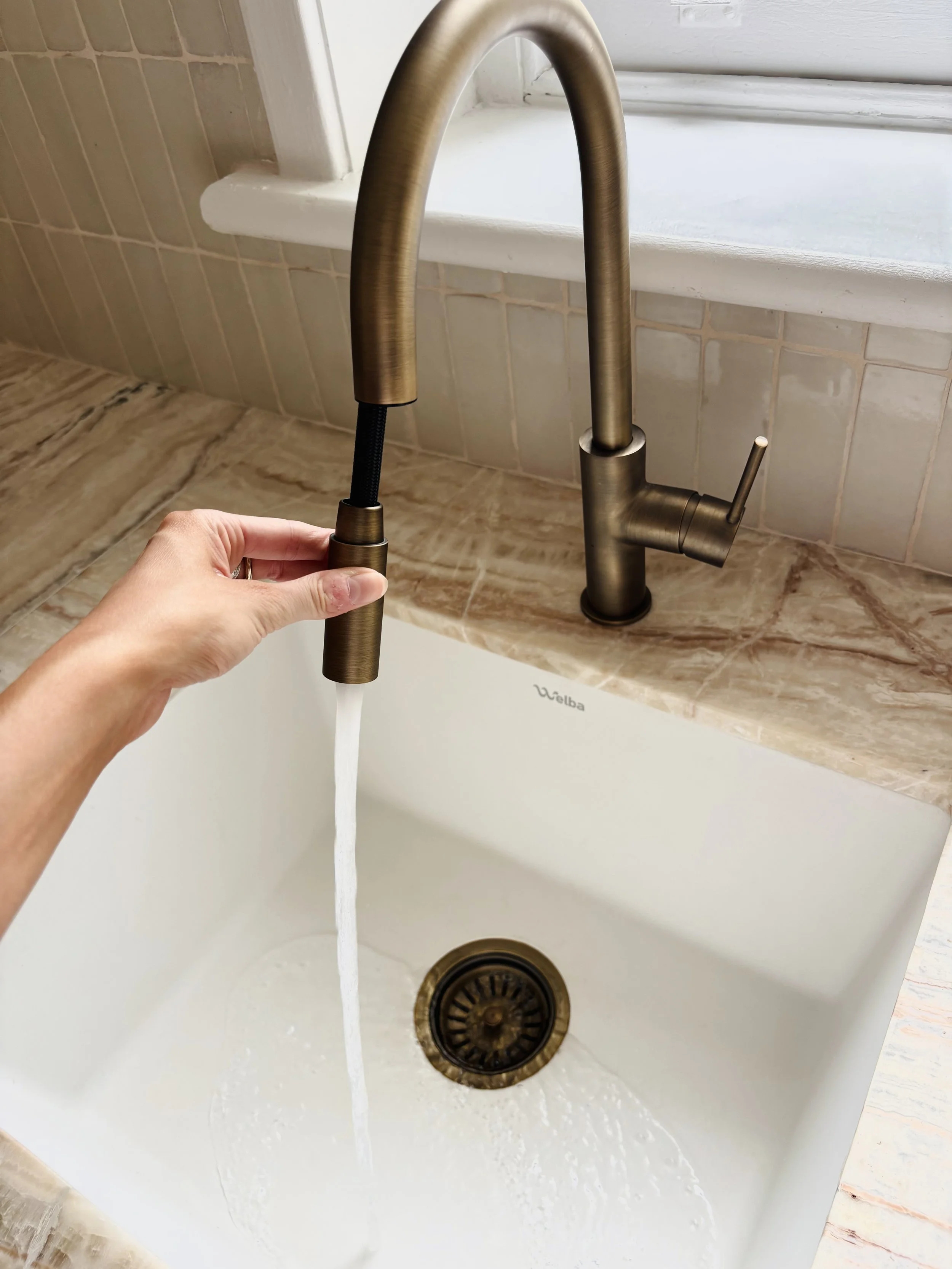 A person turning on a brushed bronze kitchen faucet with running water over a white farmhouse sink, with a beige marble countertop and a window in the background.