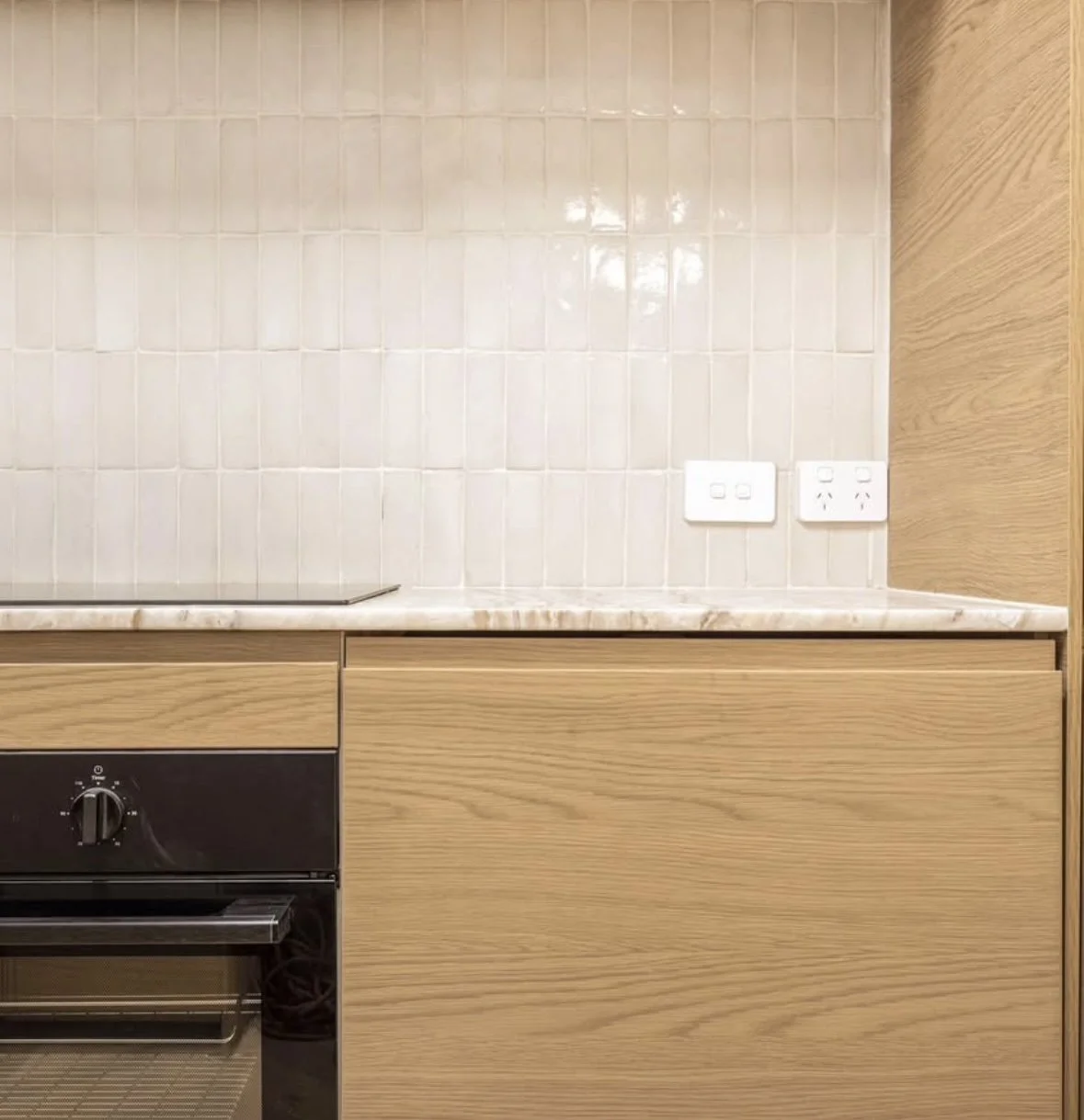 Kitchen with beige tiled wall, wooden cabinets, black oven, and two double power outlets.
