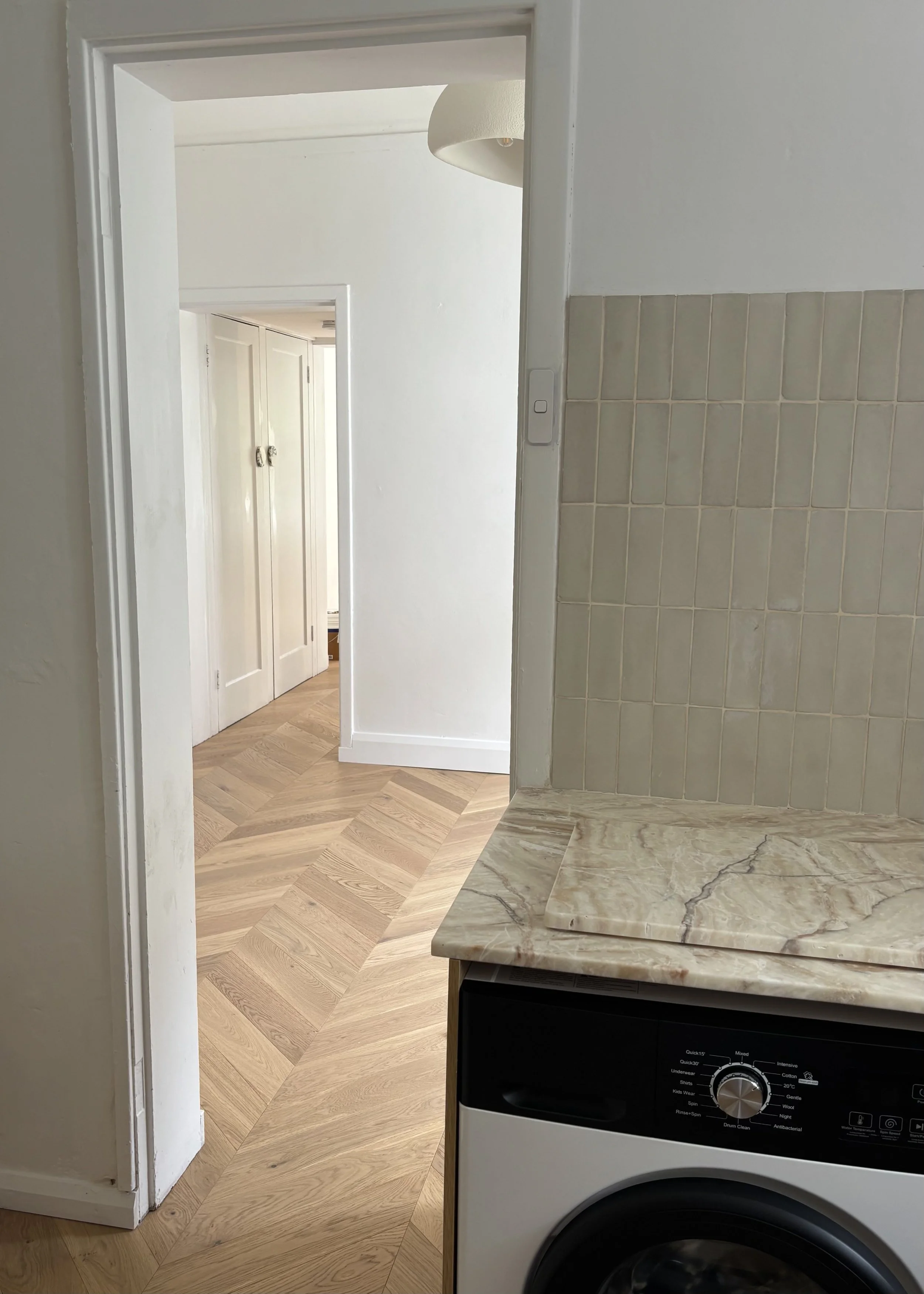 Partial view of a laundry area with a washing machine, beige tiled wall, and marble countertop, with a doorway leading to a hallway with wooden chevron flooring and white walls.