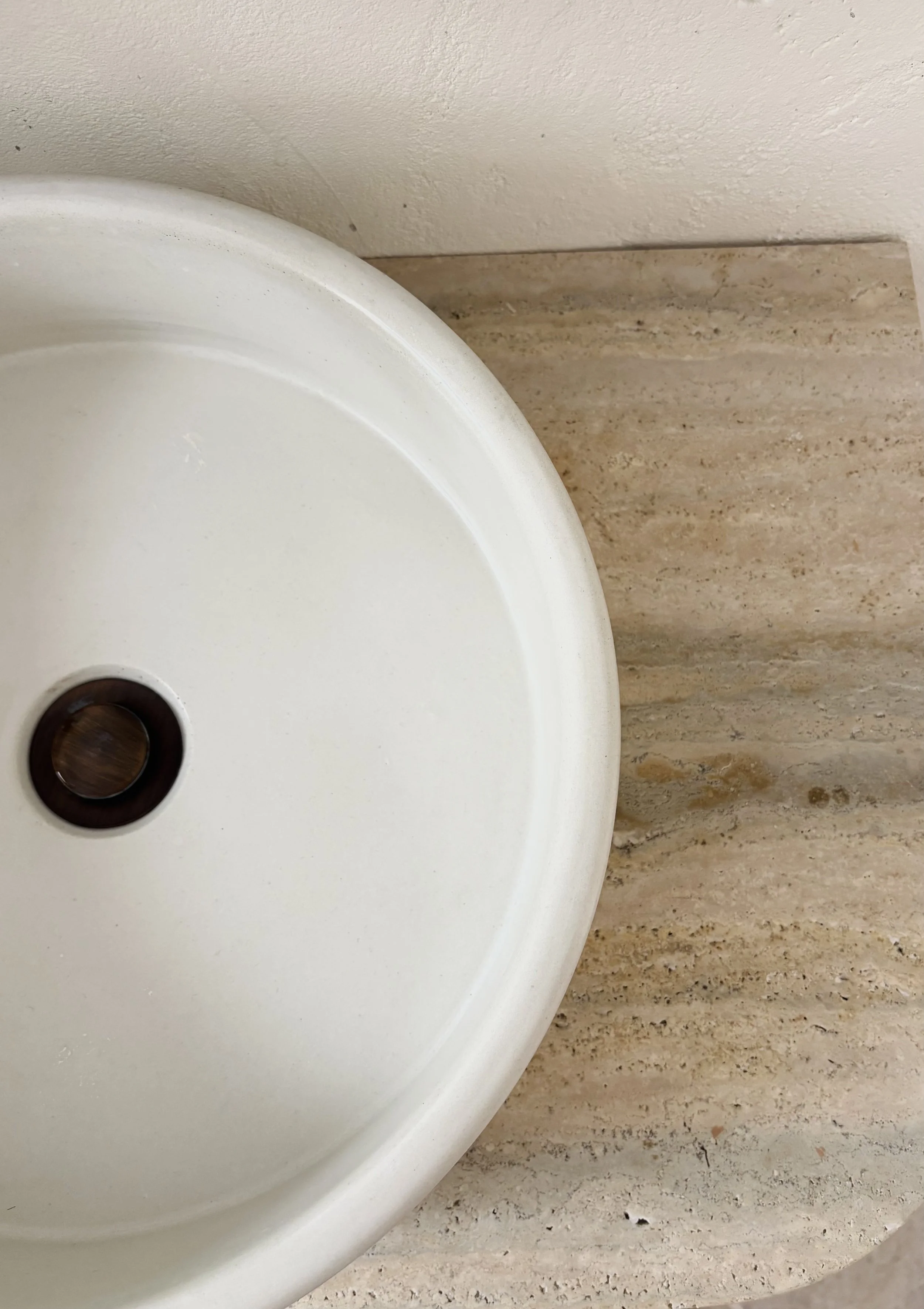 Top-down view of a white bathroom sink with a drain at the center, mounted on a beige marble countertop.