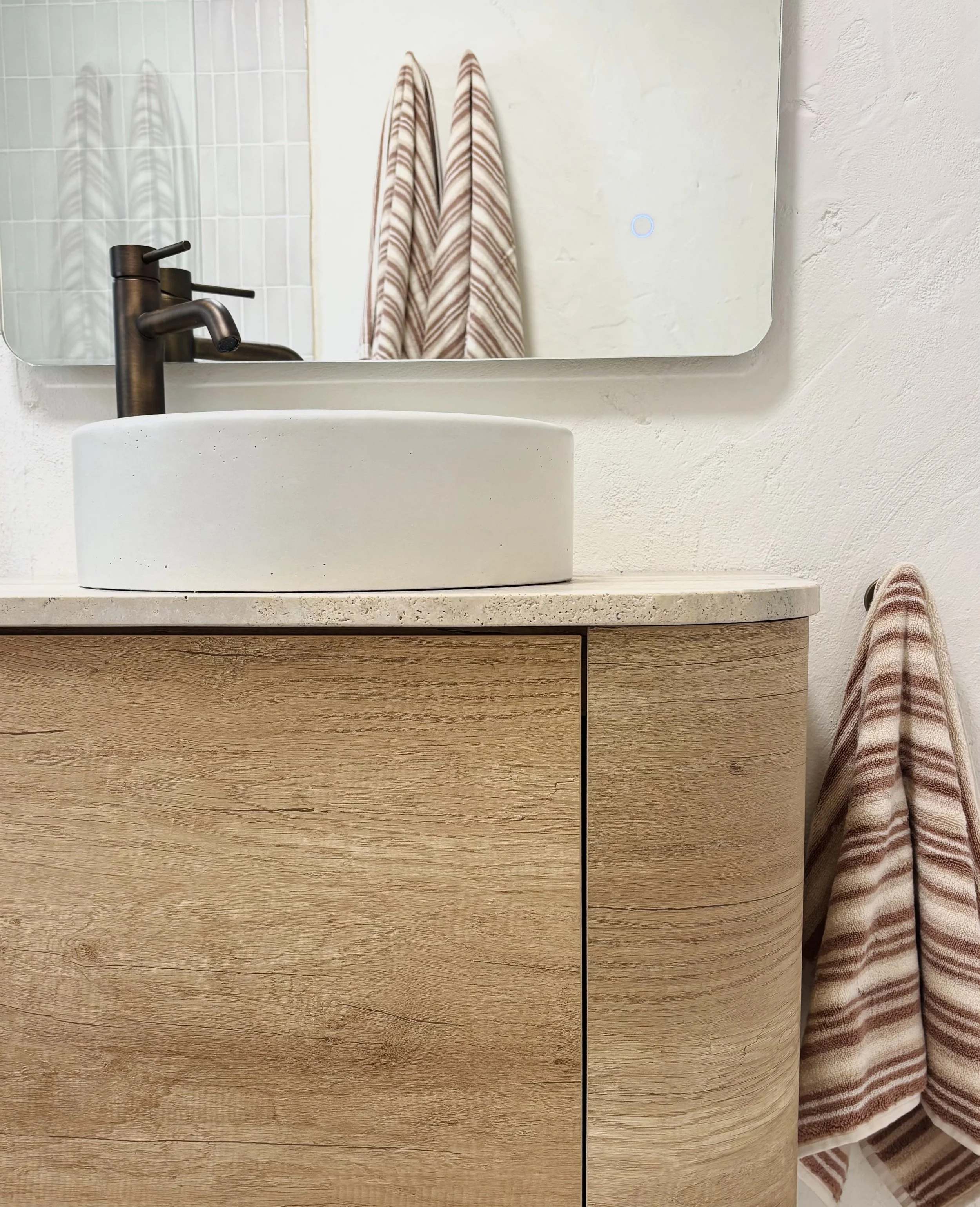 Close-up of a bathroom vanity with a round white vessel sink, a black faucet, a mirror, a wooden cabinet, and striped towels hanging.