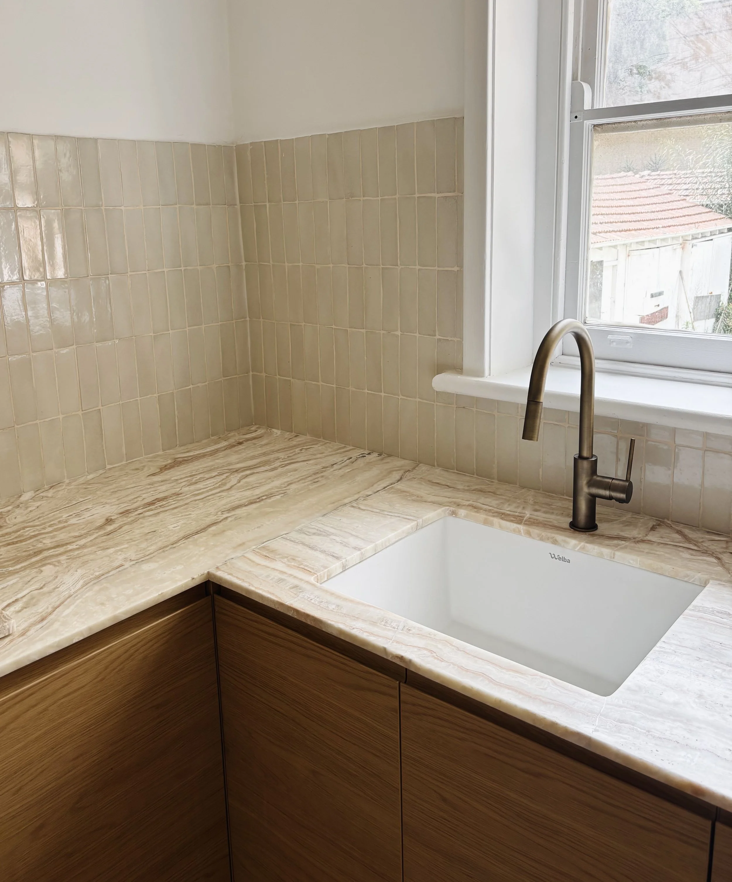 Kitchen countertop with beige marble surface, a white sink, and a brass faucet. There is a window above the sink with a view of neighboring houses.