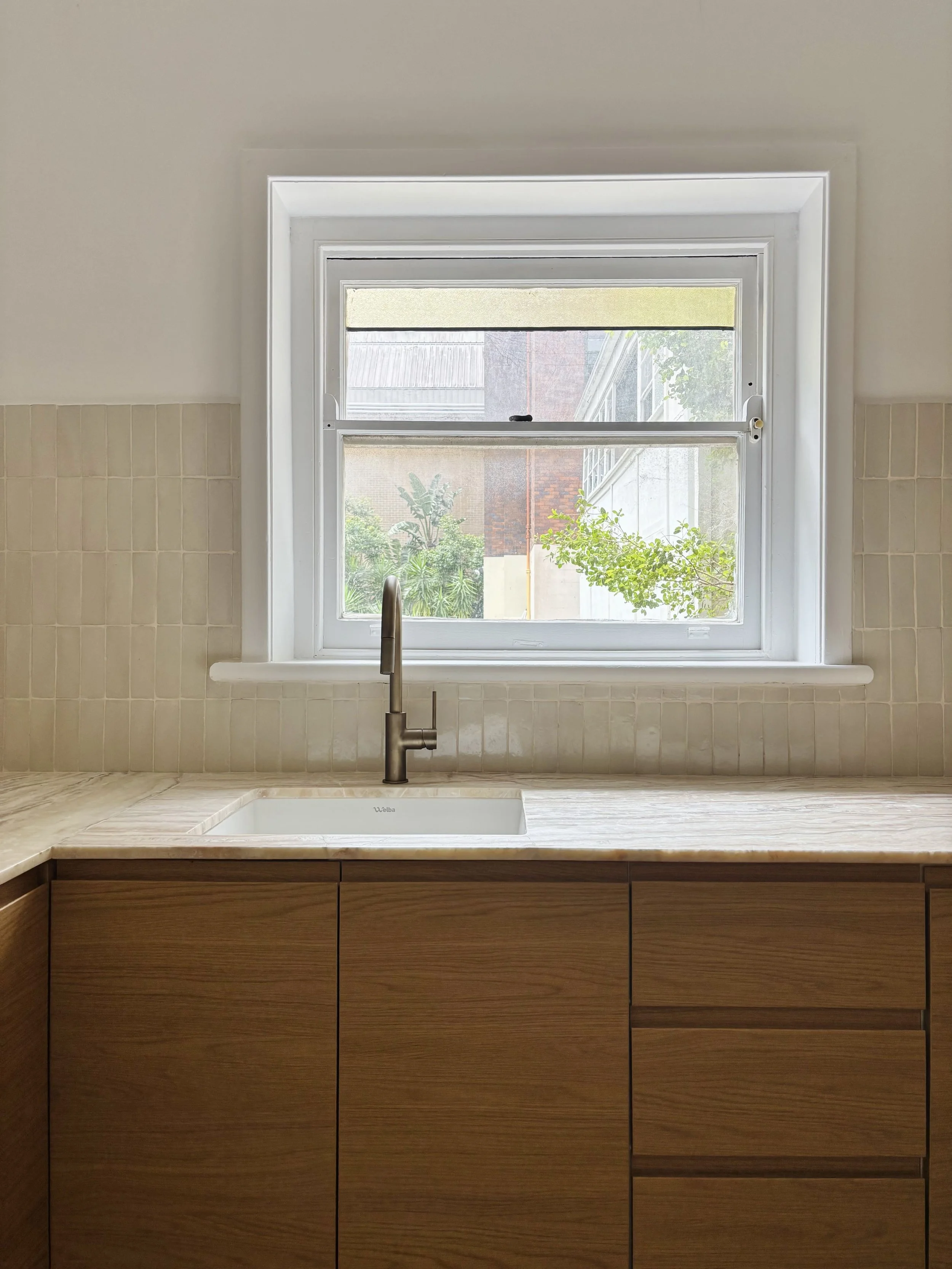 Kitchen sink under a window with a view of trees and neighboring buildings.