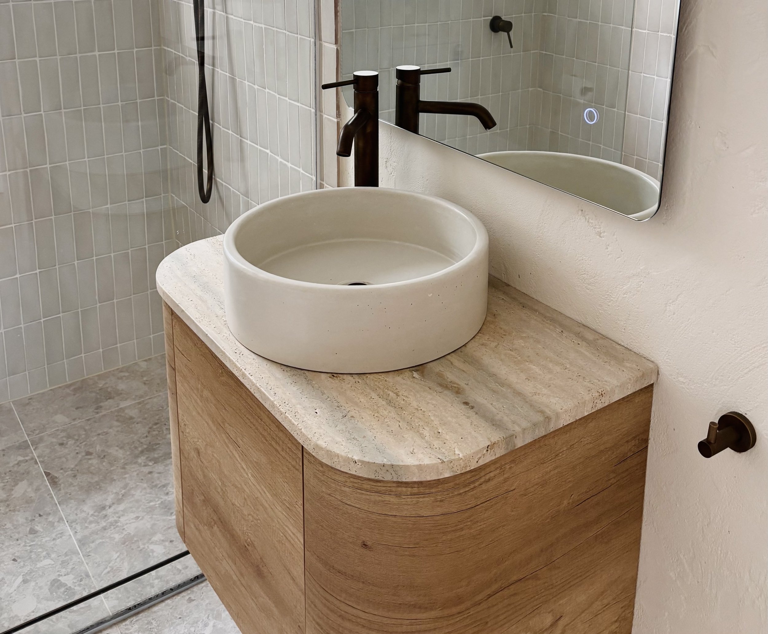 A modern bathroom vanity with a round, white vessel sink and a dark bronze faucet, a large mirror above, and a marble countertop with wooden cabinetry below.