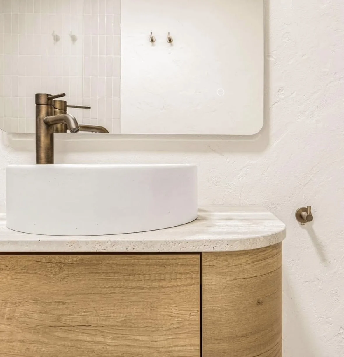 A modern bathroom vanity with a white oval vessel sink and a brushed bronze faucet, set on a light-colored marble countertop with a wooden cabinet below. A wall mirror is above the sink, and a small wall-mounted fixture is on the right side.