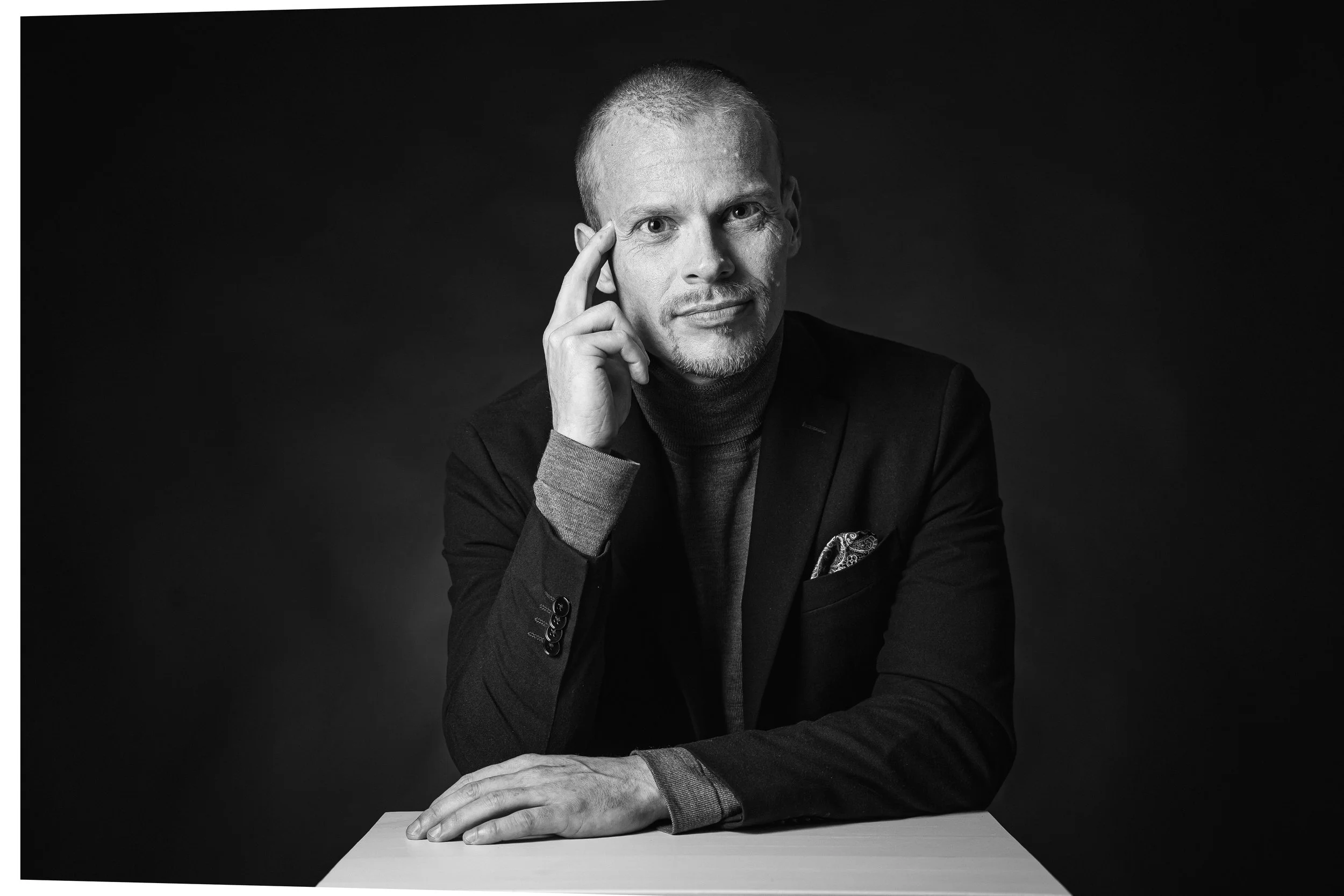 Black and white portrait of a man with short hair, wearing a dark blazer and turtleneck, sitting at a table with a plain background, looking directly at the camera with his right hand touching his temple.