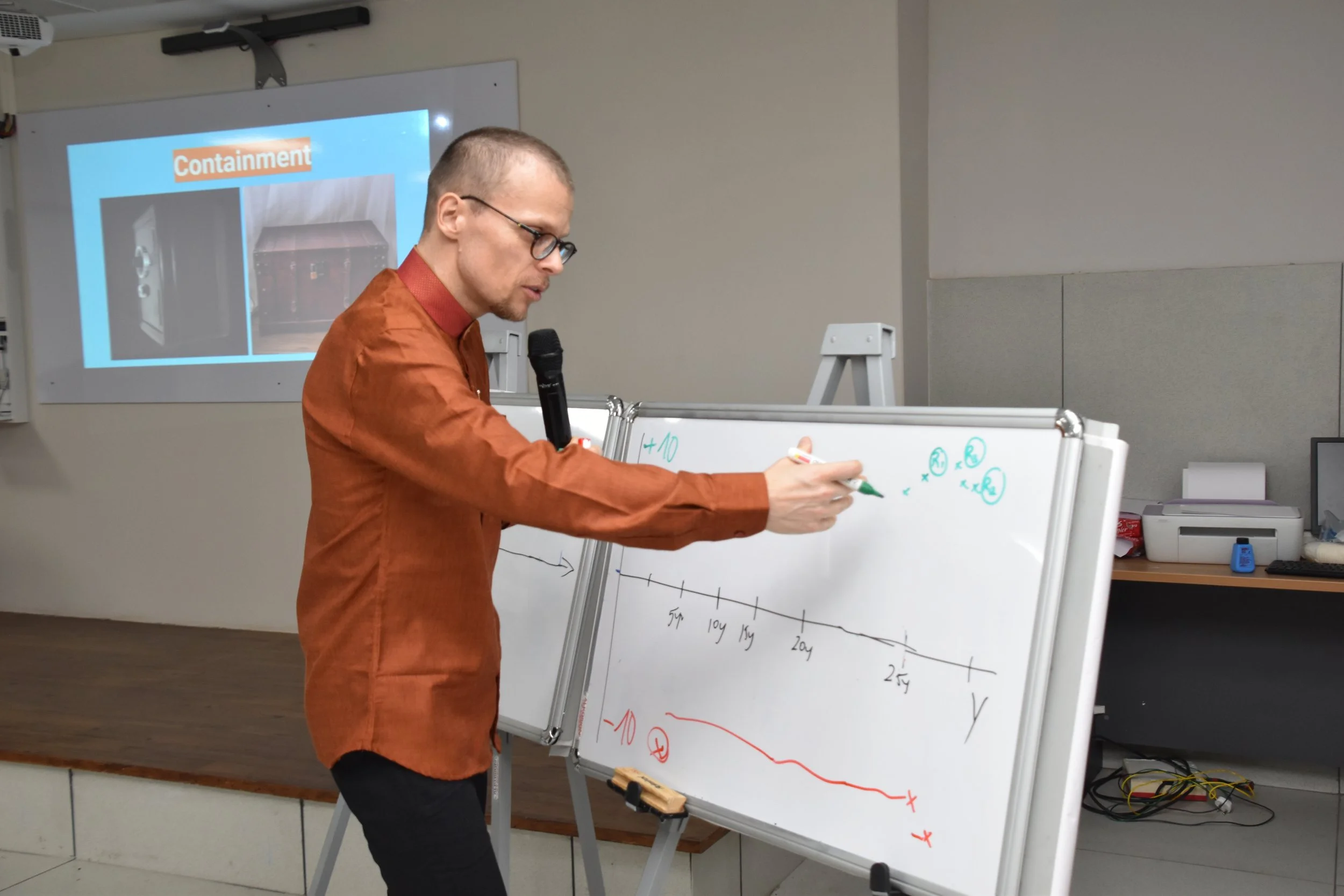 Man in orange shirt giving a presentation with whiteboard, pointing at drawings and notes, microphone in his hand, projector screen in background displaying slides about containment.