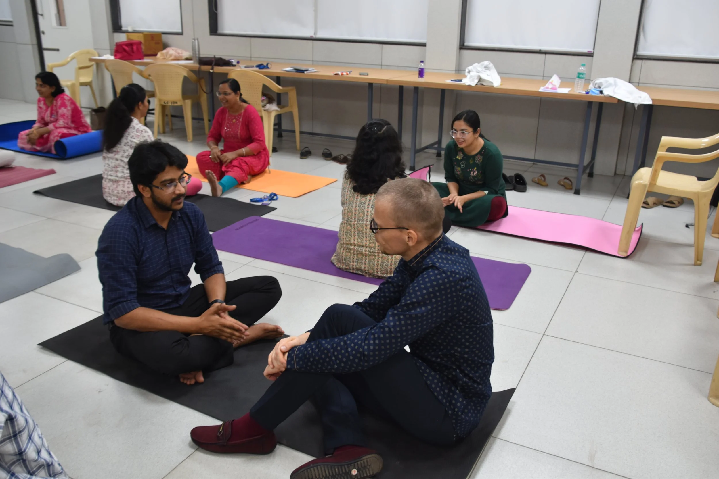 Group of people participating in a yoga or meditation session in a room, with some seated on yoga mats and engaging in conversation.