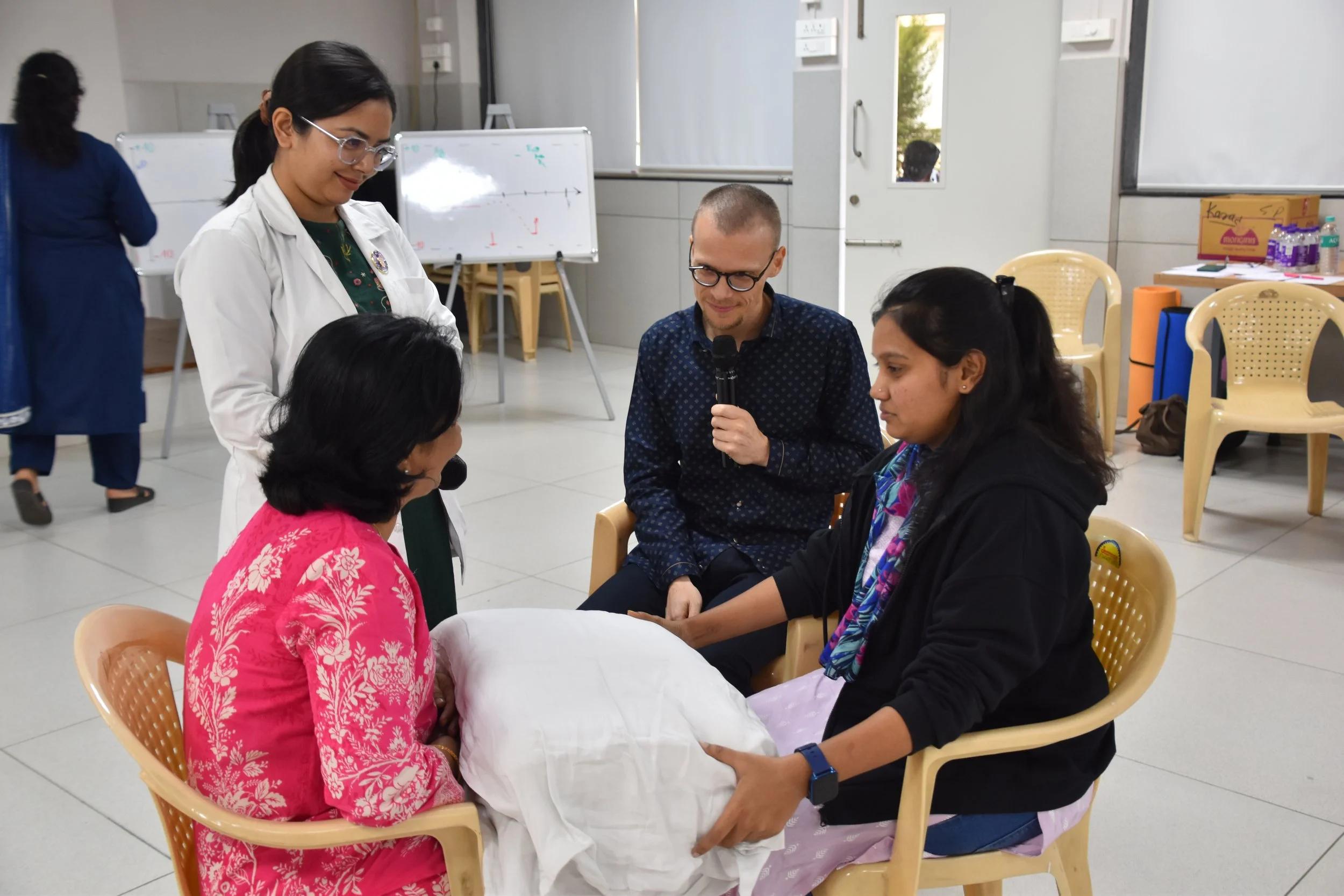 A group of four people, three women and one man, sitting and standing around a woman in a wheelchair, participating in a medical training or workshop in a classroom setting. The woman in the wheelchair is holding a pillow on her lap, and the woman st